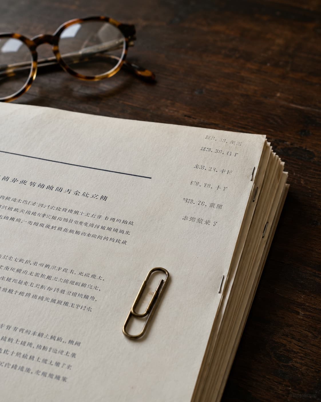 A stack of stapled cream-coloured documents with a brass paper clip on top and a pair of round-frame reading glasses resting beside on a dark walnut writing desk.