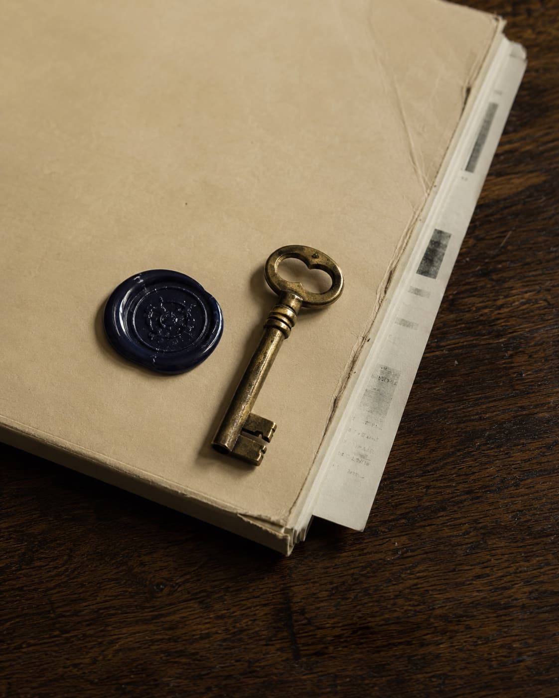 A closed manila document folder with a navy wax seal and an antique brass key resting beside it on a dark walnut writing desk.